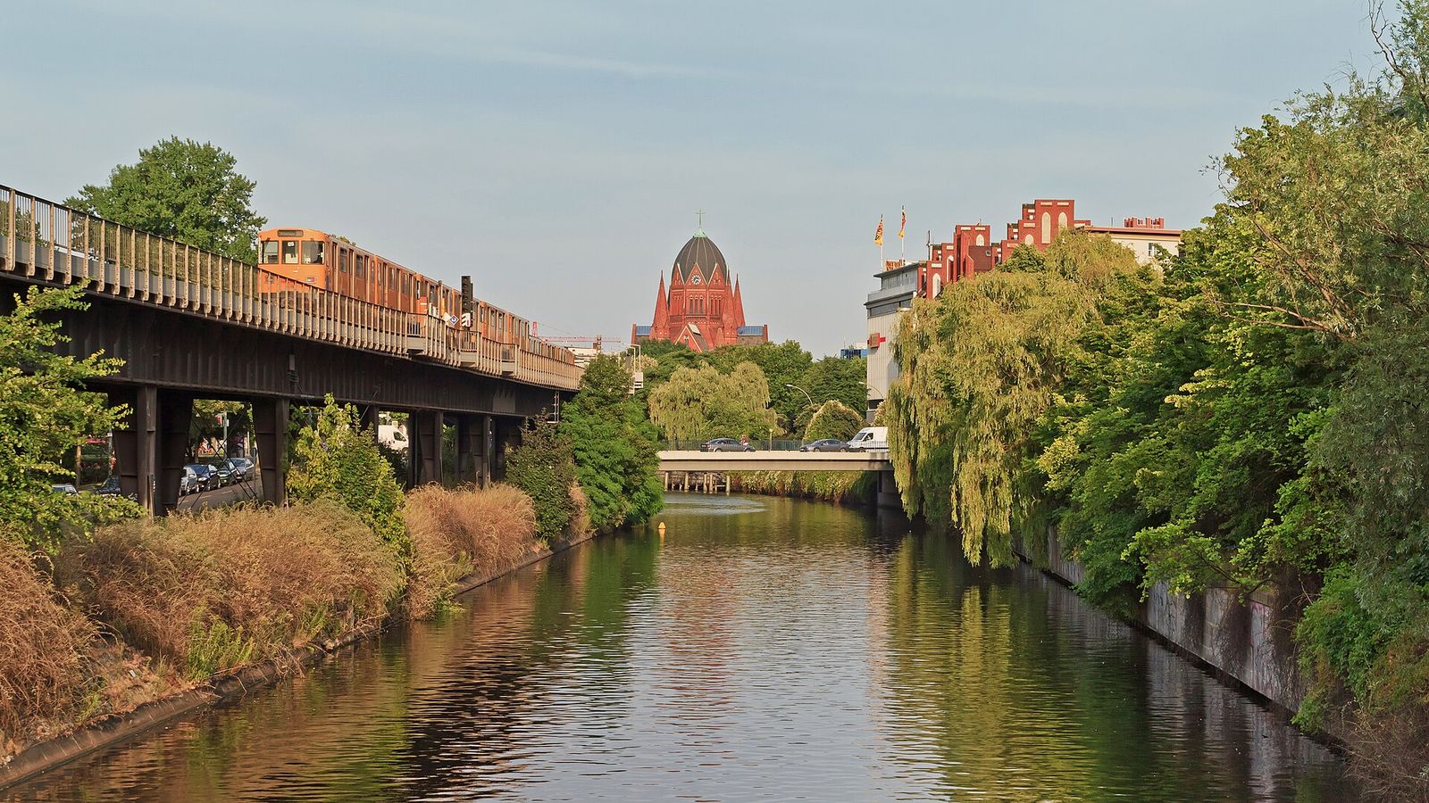 Der Landwehrkanal in Kreuzberg, links die Hochbahn auf ihrem Viadukt, rechts Trauerweiden am Ufer und eine rote Backsteinkirche im Hintergrund